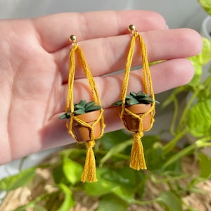 Hand holding a pair of dangling earrings that are miniature potted plants hanging from macramé. earring posts are gold. Macramé color is mustard. Pot and plant are made of polymer clay. The Pot is terracotta and the plant is green aloe.