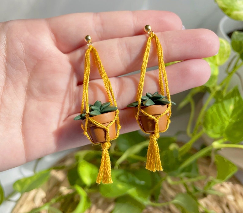 Hand holding a pair of dangling earrings that are miniature potted plants hanging from macramé. earring posts are gold. Macramé color is mustard. Pot and plant are made of polymer clay. The Pot is terracotta and the plant is green aloe.
