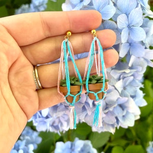 Op de afbeelding: Een paar handgemaakte oorbellen met miniatuur potplanten die hangen aan een macramé touw. De oorbellen zijn gemaakt van roze en blauw touw en hebben gouden haakjes.