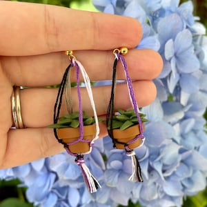Puede incluir: Un par de pendientes con suculentas en miniatura en macetas que cuelgan de cordones de estilo macramé con hilos negros, blancos, morados y marrones. Los pendientes están hechos de arcilla polimérica y son un accesorio único y caprichoso.