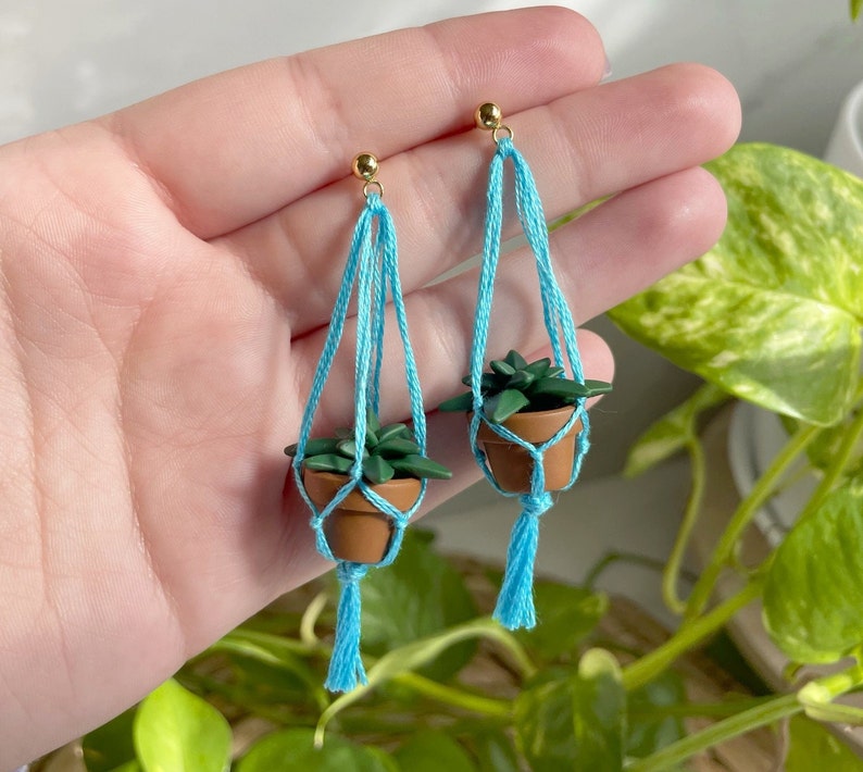 Hand holding a pair of dangling earrings that are miniature potted plants hanging from macramé. Earring posts are gold. Macramé color is aqua. Pot and plant are made of polymer clay. The Pot is terracotta and the plant is green aloe.