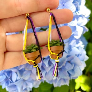 May include: A pair of earrings featuring miniature potted plants hanging from macrame-style cords with yellow, white, and purple threads. The earrings are made with gold-colored metal hooks.