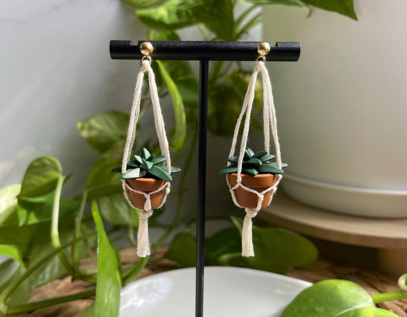 A pair of dangling earrings that are miniature potted plants hanging from macramé. Displayed on black dangle earring display. Earring posts are gold. Macramé color is natural/off white. Pot and plant are made of polymer clay.