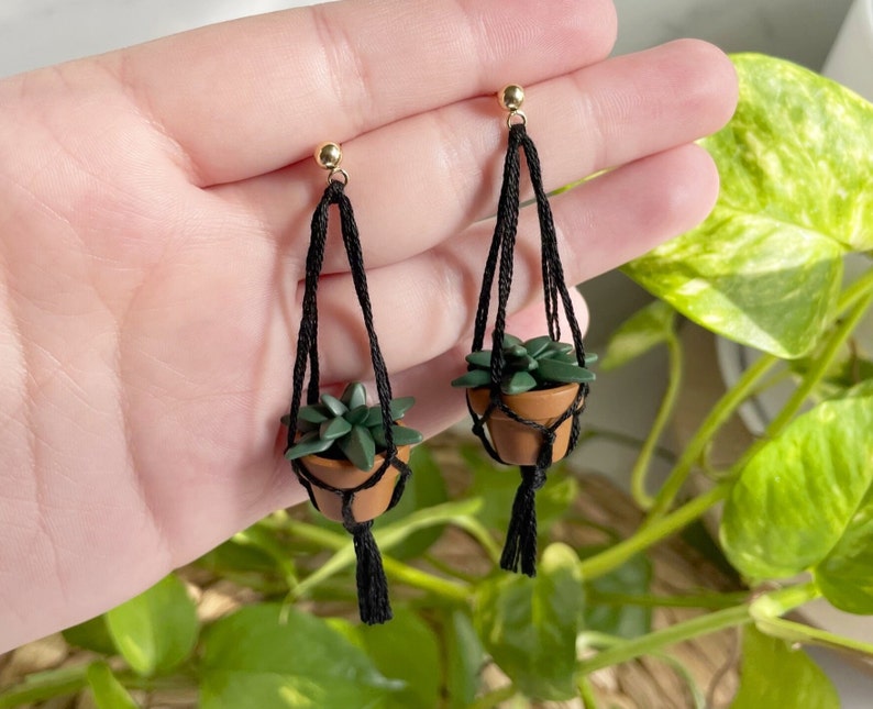 Hand holding a pair of dangling earrings that are miniature potted plants hanging from macramé. Earring posts are gold. Macramé color is black. Pot and plant are made of polymer clay. The Pot is terracotta and the plant is green aloe.