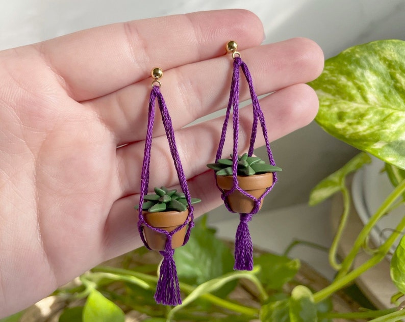 Hand holding a pair of dangling earrings that are miniature potted plants hanging from macramé. Earring posts are gold. Macramé color is grape. Pot and plant are made of polymer clay. The Pot is terracotta and the plant is green aloe.