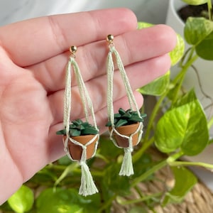 Hand holding a pair of dangling earrings that are miniature potted plants hanging from macramé. Earring posts are gold. Macramé color is seafoam green. Pot and plant are made of polymer clay. The Pot is terracotta and the plant is green aloe.