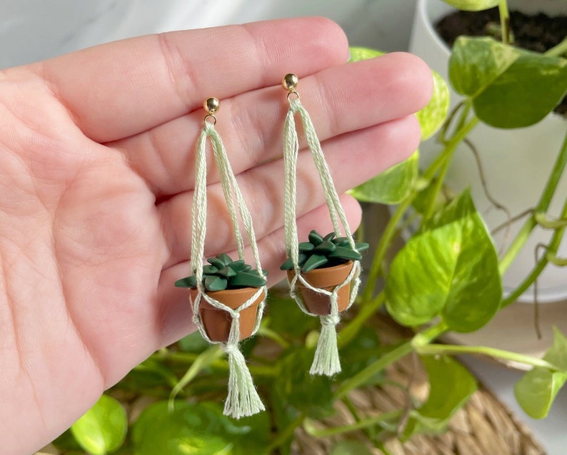 Hand holding a pair of dangling earrings that are miniature potted plants hanging from macramé. Earring posts are gold. Macramé color is seafoam green. Pot and plant are made of polymer clay. The Pot is terracotta and the plant is green aloe.