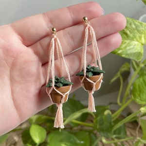 Hand holding a pair of dangling earrings that are miniature potted plants hanging from macramé. Earring posts are gold. Macramé color is peach. Pot and plant are made of polymer clay. The Pot is terracotta and the plant is green aloe.