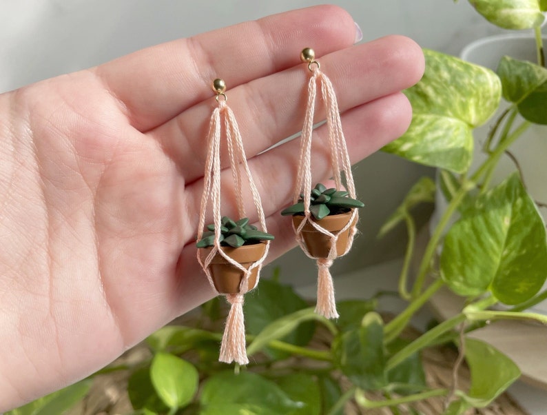 Hand holding a pair of dangling earrings that are miniature potted plants hanging from macramé. Earring posts are gold. Macramé color is peach. Pot and plant are made of polymer clay. The Pot is terracotta and the plant is green aloe.