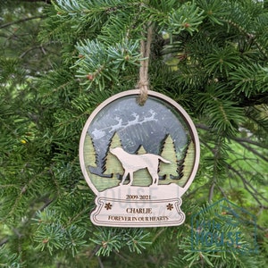 May include: A wooden Christmas bauble with a snow globe design. The globe features a silhouette of a dog in a winter forest scene with Santa's sleigh flying overhead. The bauble is personalised with the text "2009-2021 Charlie Forever in Our Hearts".