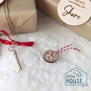 以下が含まれることがあります： 「Property of the North Pole」と書かれた木製のボタンのオーナメントと赤と白の紐。オーナメントは白いふわふわの表面の上に置かれています。