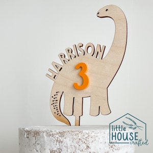 Pode incluir: Topper de bolo de dinossauro de madeira com o nome "HARRISON" e o n&uacute;mero "3" em laranja. O topper est&aacute; em um bolo branco coberto com glac&ecirc;.