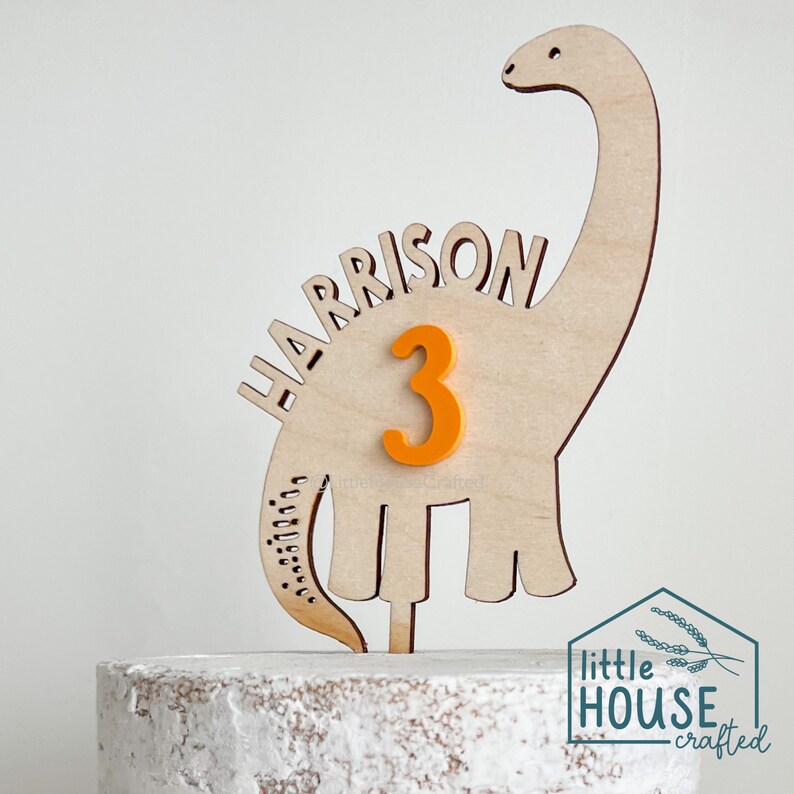 Pode incluir: Topper de bolo de dinossauro de madeira com o nome "HARRISON" e o n&uacute;mero "3" em laranja. O topper est&aacute; em um bolo branco coberto com glac&ecirc;.