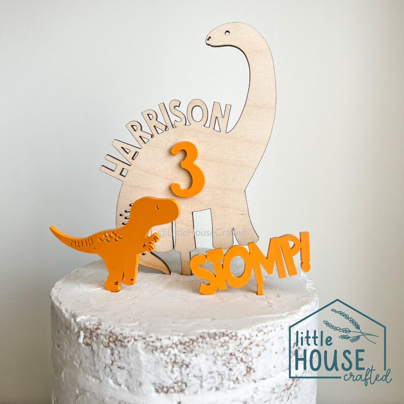 Pode incluir: Um conjunto de topo de bolo de dinossauro de madeira com um dinossauro castanho, um dinossauro laranja mais pequeno, o n&uacute;mero 3 e a palavra "STOMP!" em laranja. O conjunto est&aacute; sobre um bolo branco coberto com glac&ecirc; com o nome "HARRISON" escrito na lateral.
