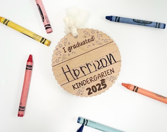 May include: A wooden graduation ornament with the text "I graduated! Harrison Kindergarten 2025" surrounded by colorful crayons. The ornament is round with a scalloped edge and a white ribbon.