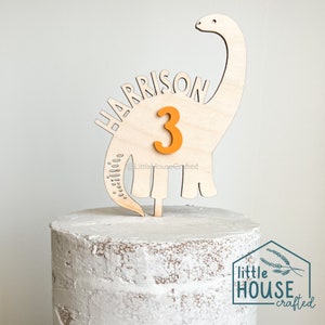Pode incluir: Topper de bolo de dinossauro de madeira com o nome Harrison e o n&uacute;mero 3 em laranja. O dinossauro &eacute; marrom claro e tem um pesco&ccedil;o e uma cauda longos. O topper est&aacute; em um bolo branco.