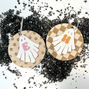 Könnte beinhalten: Zwei geisterförmige Schlüsselanhänger aus Holz mit einem karierten und floralen Muster. Die Geister haben einen weißen Umriss und ein pinkfarbenes "E" und ein orangefarbenes "H" im Inneren.