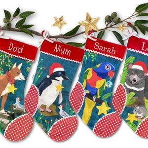 May include: Four Christmas stockings, each with a name: "Dad", "Mum", "Sarah", and "Leo". The stockings feature illustrations of a dingo, penguin, parrot, and Tasmanian devil, all wearing Santa hats. The stockings are decorated with red and white polka dots, with a sprig of leaves above.