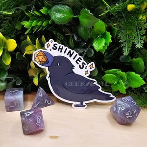 May include: A black crow with a blue beak holding a gold gemstone in its beak. The crow is holding a sign that says "Shinies". The crow is surrounded by purple and white dice.