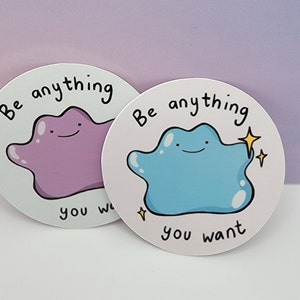 May include: Two round stickers with a white background and black text that reads "Be anything you want". Each sticker features a cartoon purple and blue blob-like creature with a happy expression.