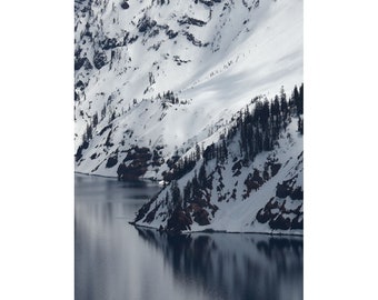 May include: A snowy mountainside with a dark blue lake at the base. The mountain is covered in snow and ice, with a few trees growing on the slopes. The lake is calm and reflects the sky.