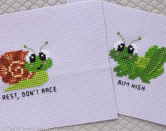 May include: Two cross-stitch embroidery patterns featuring cute cartoon animals. The first pattern shows a snail with the text "Rest, Don't Race". The second pattern shows a caterpillar with the text "Aim High".
