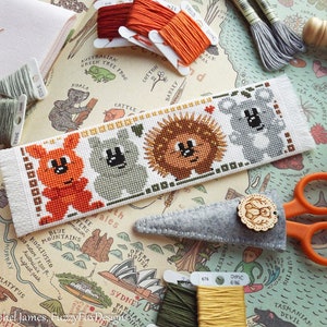 May include: A white bookmark with a cross-stitch design featuring four Australian animals: a red kangaroo, a gray koala, a brown echidna, and a gray koala. The bookmark is surrounded by embroidery floss and a map of Australia.