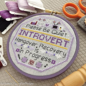 May include: A purple embroidery hoop with a cross-stitch design that reads "Please Be Quiet Introvert Hangover. Recovery is in Progress".