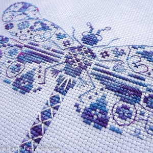 K&ouml;nnte beinhalten: Nahaufnahme einer Kreuzstich-Stickerei einer Libelle mit filigranen Details in Blaut&ouml;nen, Lila und Wei&szlig; auf einem wei&szlig;en Aida-Stoff.