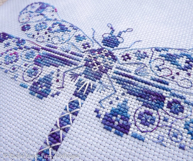 K&ouml;nnte beinhalten: Nahaufnahme einer Kreuzstich-Stickerei einer Libelle mit filigranen Details in Blaut&ouml;nen, Lila und Wei&szlig; auf einem wei&szlig;en Aida-Stoff.