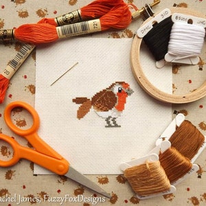 May include: A cross-stitch project featuring a robin with orange, brown, and white details. The image includes orange scissors, embroidery floss, a needle, and a wooden embroidery hoop with black and white thread. The project is on a white fabric.