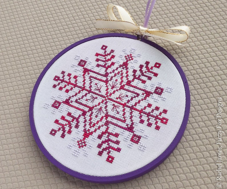 May include: A purple and white snowflake cross-stitch ornament with a white ribbon.