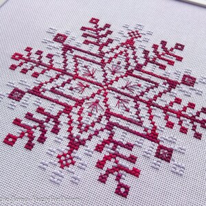 May include: A cross-stitch pattern of a snowflake in shades of pink and purple on white Aida cloth.
