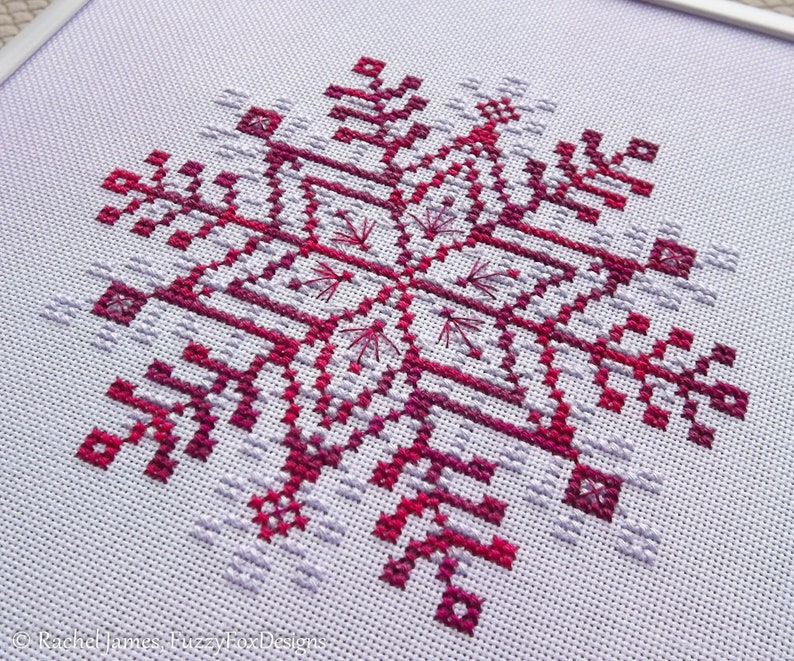 May include: A cross-stitch pattern of a snowflake in shades of pink and purple on white Aida cloth.