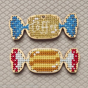 May include: Two wooden candy-shaped ornaments with cross-stitch designs. The top ornament is yellow with gold glitter and the words "Tootsie" stitched in white. The bottom ornament is yellow and brown with a white cross-stitch design.