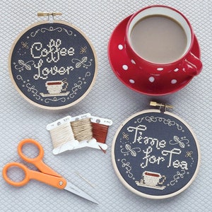 May include: Two embroidery hoops with black fabric and white embroidery. The top hoop says "Coffee Lover" and the bottom hoop says "Time for Tea". Both hoops have a small cup of coffee or tea embroidered in the centre.