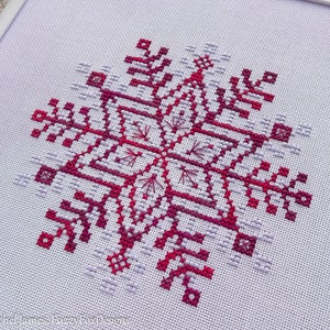 May include: A close-up of a cross-stitch pattern of a snowflake in shades of pink and purple on white Aida cloth.