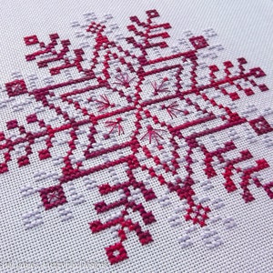 May include: A close-up of a cross-stitch pattern of a snowflake in shades of pink and red on a white background.