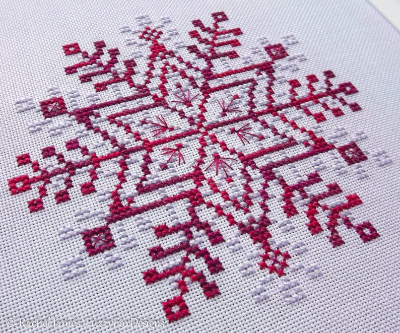 May include: A close-up of a cross-stitch pattern of a snowflake in shades of pink and red on a white background.