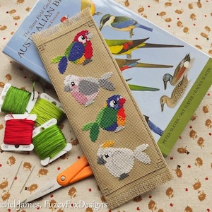 May include: A cross-stitch bookmark featuring five colorful birds. The birds are a cockatoo, a parrot, a lorikeet, a galah, and a budgie. The bookmark is on a beige background with a fringe border.