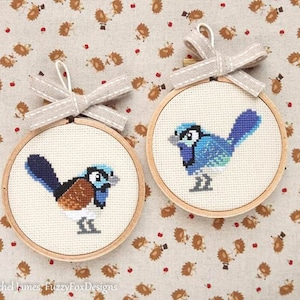 May include: Two cross-stitch bird ornaments. Each bird is in a wooden hoop, with a light beige background. The birds are blue, brown, and white. Each ornament has a beige ribbon bow at the top.
