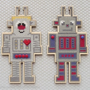 May include: Two wooden robot ornaments with red and blue details. The robot on the left has a yellow antenna and a red heart on its chest. The robot on the right has a blue antenna and a blue button on its chest.