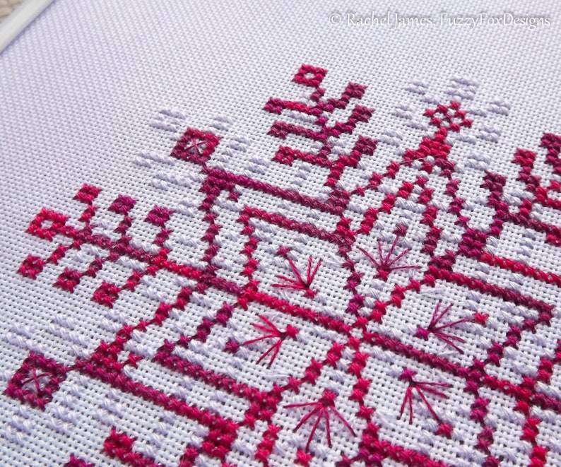 May include: Close-up of a cross-stitch pattern with a geometric design in shades of pink and purple on white Aida cloth.