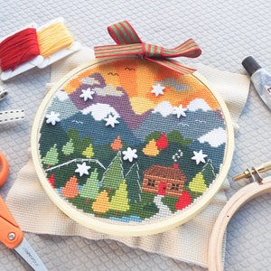 K&ouml;nnte beinhalten: Ein fertiger Kreuzstich-Stickrahmen mit einer Winterszene mit einer H&uuml;tte im Wald. Der Rahmen ist von Bastelutensilien umgeben, darunter Stickgarn, Schere und Kleber.
