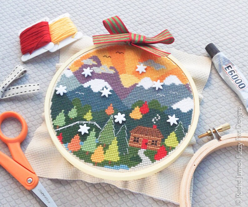 K&ouml;nnte beinhalten: Ein fertiger Kreuzstich-Stickrahmen mit einer Winterszene mit einer H&uuml;tte im Wald. Der Rahmen ist von Bastelutensilien umgeben, darunter Stickgarn, Schere und Kleber.