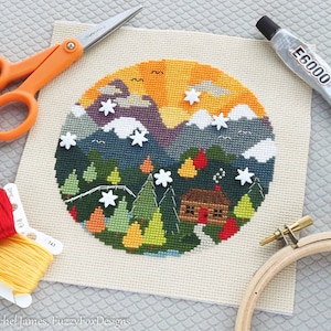 K&ouml;nnte beinhalten: Ein fertiggestelltes Kreuzstich-Stickprojekt, das eine H&uuml;tte in einer schneebedeckten Berglandschaft zeigt. Die Stickerei ist in einem Holzrahmen gespannt und von Bastelutensilien wie Stickgarn, Schere und Kleber umgeben.