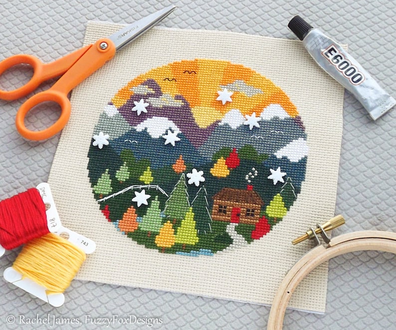 K&ouml;nnte beinhalten: Ein fertiggestelltes Kreuzstich-Stickprojekt, das eine H&uuml;tte in einer schneebedeckten Berglandschaft zeigt. Die Stickerei ist in einem Holzrahmen gespannt und von Bastelutensilien wie Stickgarn, Schere und Kleber umgeben.