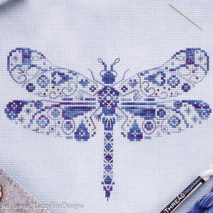 K&ouml;nnte beinhalten: Ein Kreuzstichmuster einer blauen Libelle mit filigranen Details. Die Libelle ist auf wei&szlig;em Aida-Stoff gestickt.