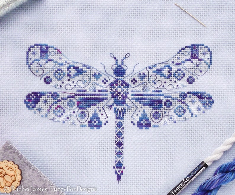 K&ouml;nnte beinhalten: Ein Kreuzstichmuster einer blauen Libelle mit filigranen Details. Die Libelle ist auf wei&szlig;em Aida-Stoff gestickt.