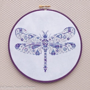 K&ouml;nnte beinhalten: Eine gestickte Libelle in Lila und Wei&szlig; mit Kreuzstich in einem runden Holzrahmen mit einem violetten Stoffr&uuml;cken.